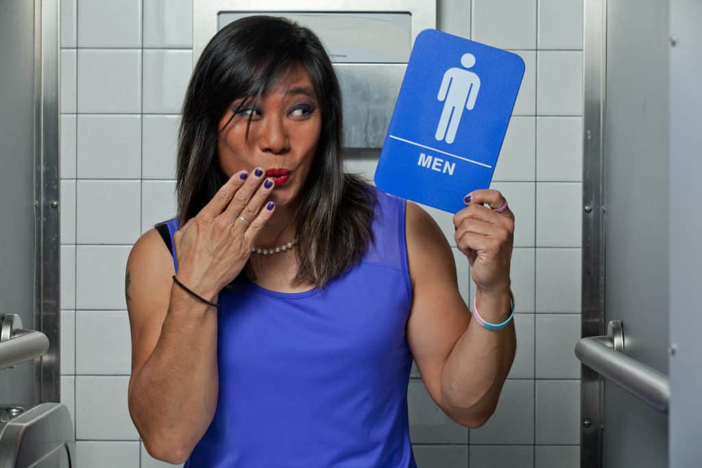 Transgender female in restroom stall with men's restroom sign in hand, surprised with fingers over mouth.