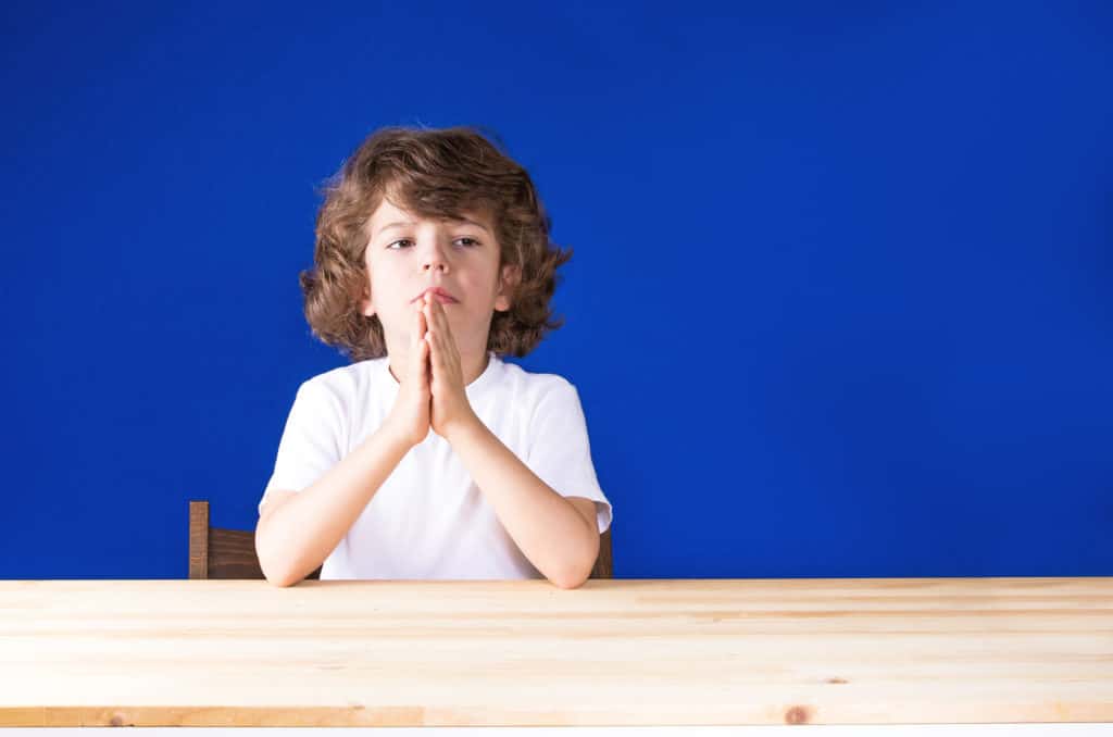 Curly cute boy clasped his hands in prayer and sad looking forward. Close-up. Blue background.