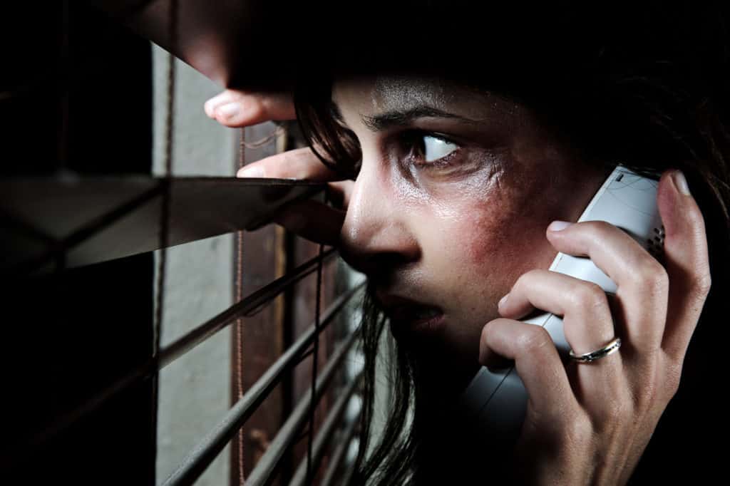 Fearful battered woman peeking through the blinds to see if her husband is home while calling for help