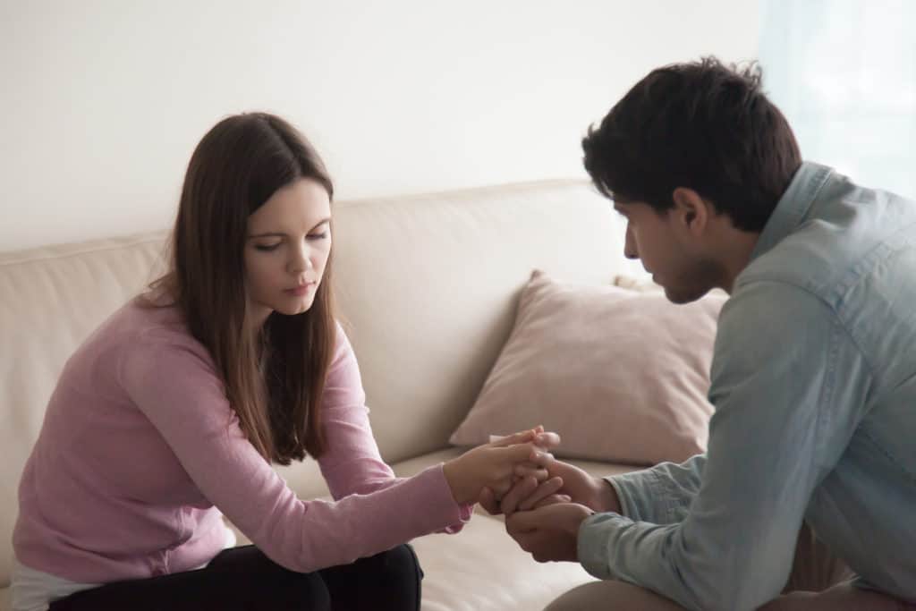 Sad couple sitting opposite one another, man holding hands of a woman. Loving husband comforting troubled young wife, compassionate boyfriend supporting crying girl, abortion or infertility problem