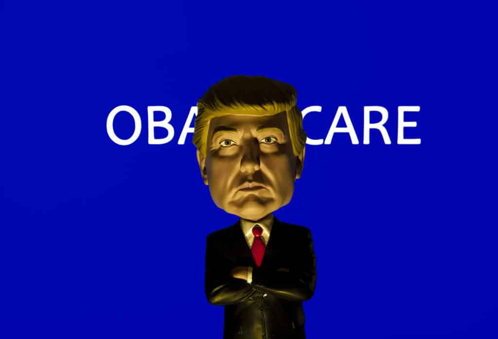 Donald Trump Caricature Bobble Head standing in front of a sign reading Obamacare - also known as the Affordable Healthcare Act