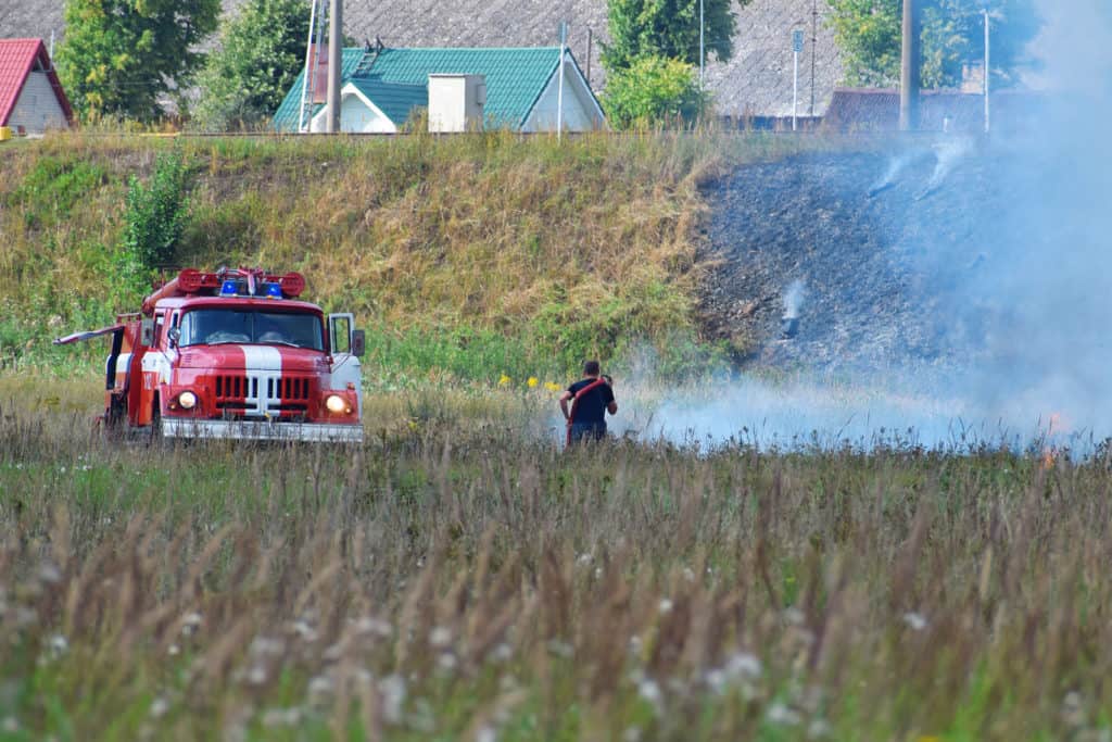 Fire truck and firefighter on firefighting rescue operation fighting with flames in burning meadow near to railway in hot summer day. Firefighting rescue operation concept.