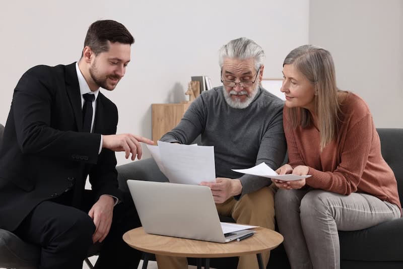 A professional attorney in a suit sits with an older couple on their couch, pointing to documents while explaining legal options
