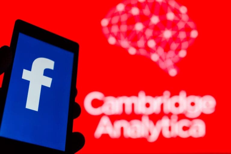 Hand holding a smartphone showing the Facebook logo, with the Cambridge Analytica name in the background