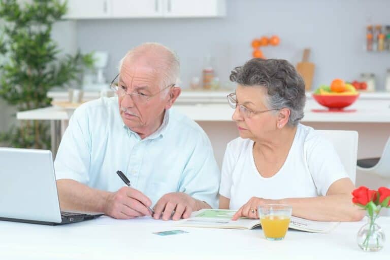 Older couple sitting at a kitchen table reviewing paperwork next to a laptop