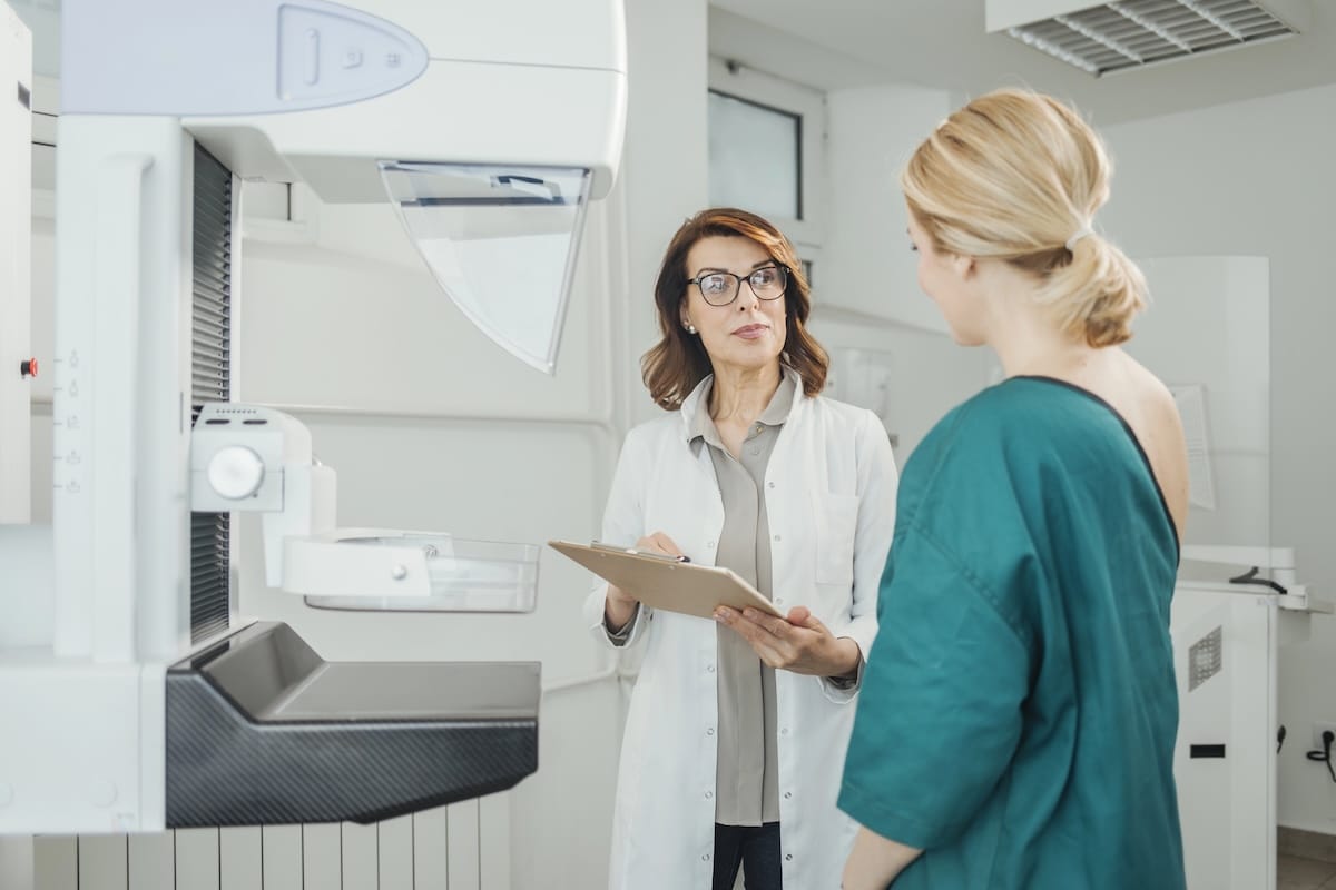 Woman in hospital gown consulting with female doctor next to mammography machine during breast health appointment