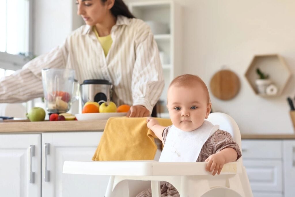 A baby sits in a high chair while a mother uses a blender to prepare baby food in the kitchen