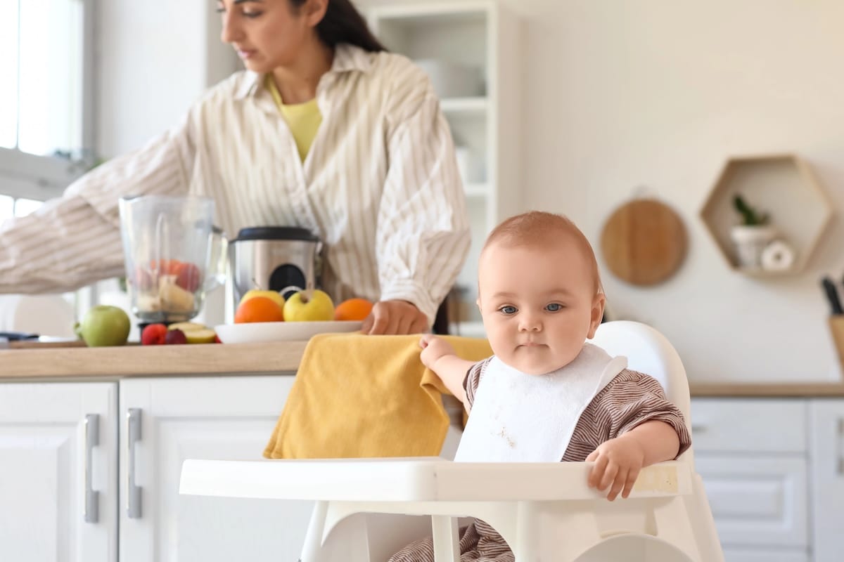 A baby sits in a high chair while a mother uses a blender to prepare baby food in the kitchen