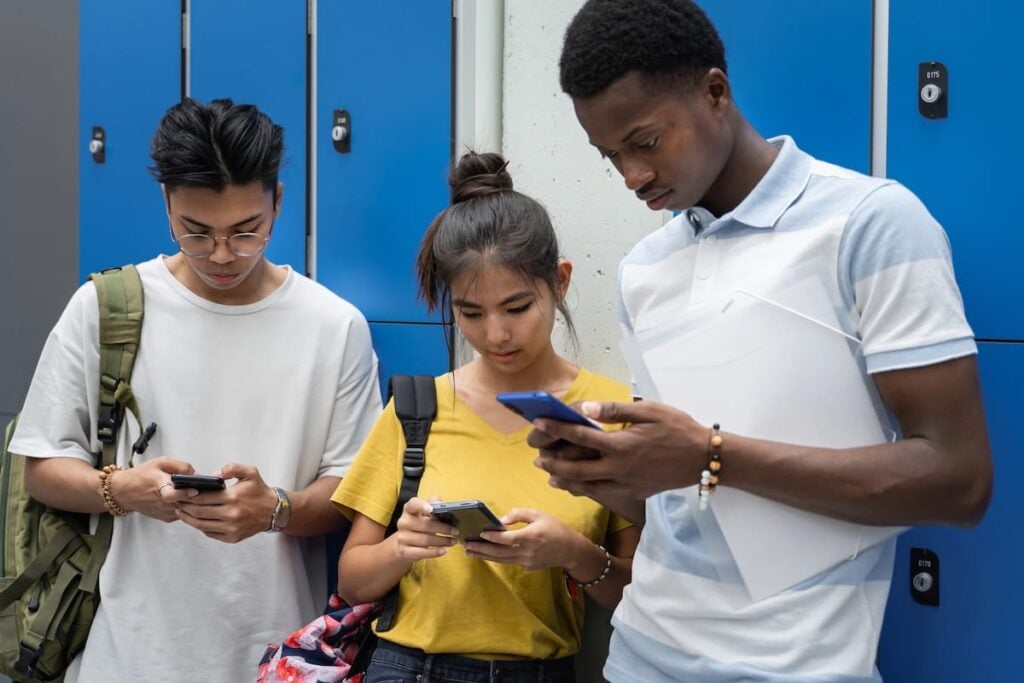 Three teenagers using smartphones at school, illustrating teen social media use and addiction concerns.
