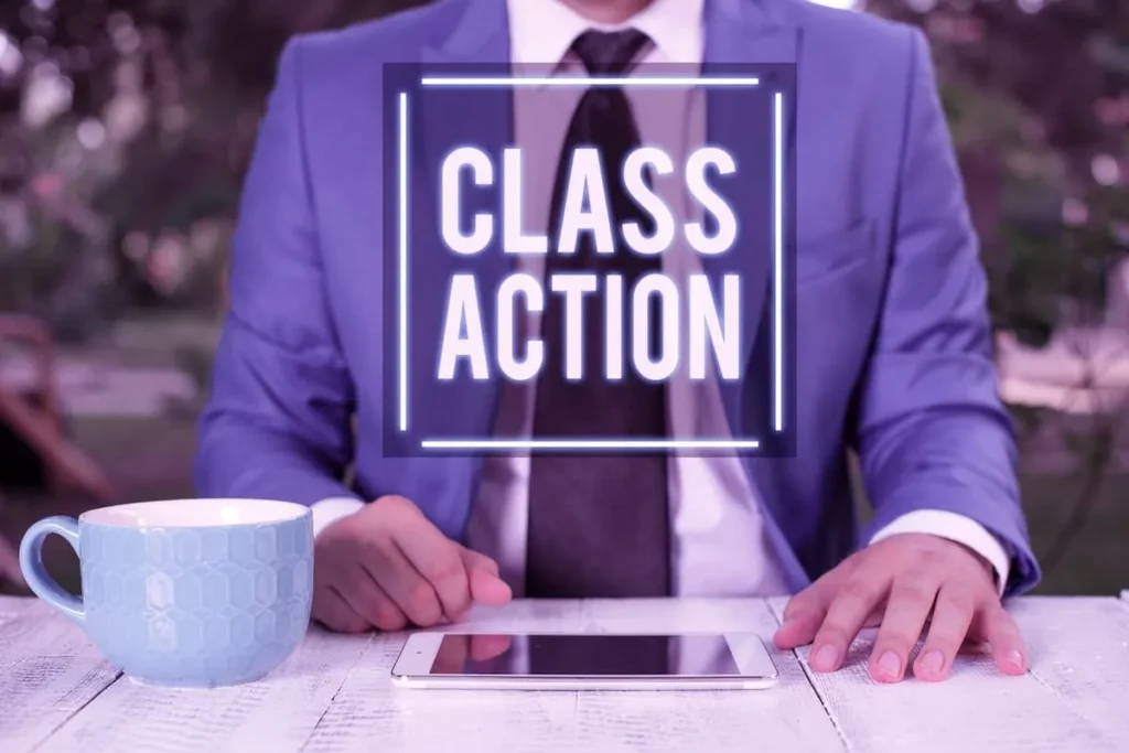 Man in a suit seated at a table behind a lit Class Action sign