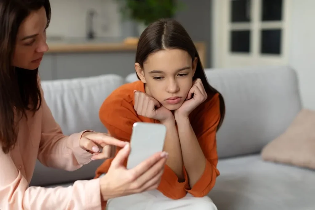 Mother holding a smartphone in front of her teenage daughter, who sits on the couch with her chin in her hands