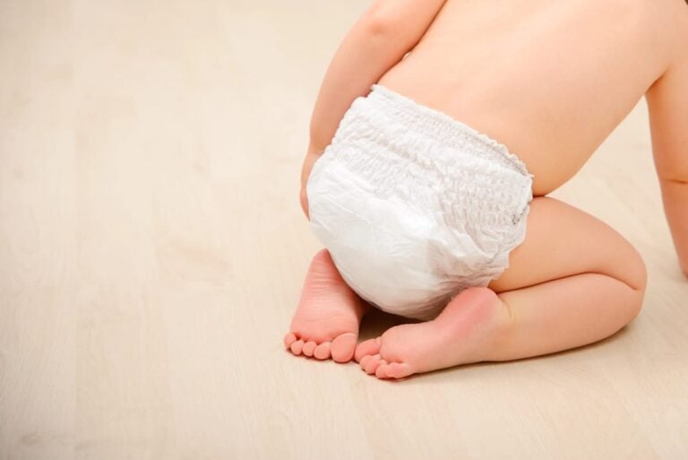 Baby wearing a disposable diaper, seen from behind while kneeling on a light wood floor