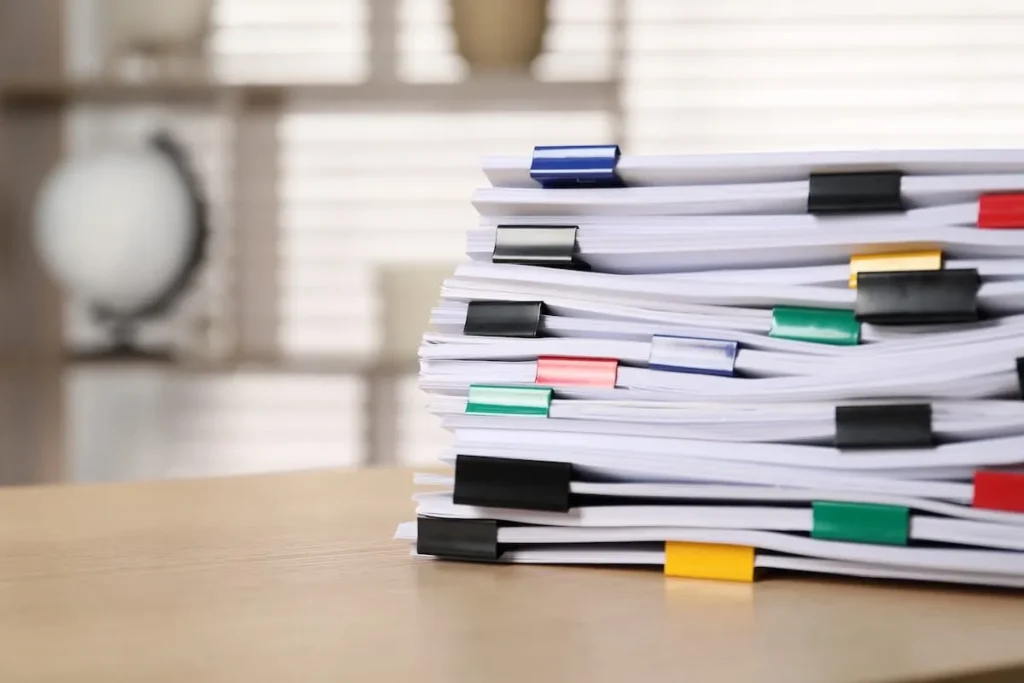 Tall stack of paper documents held together with colored binder clips on a wooden desk