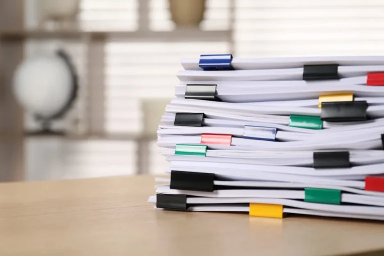 Tall stack of paper documents held together with colored binder clips on a wooden desk