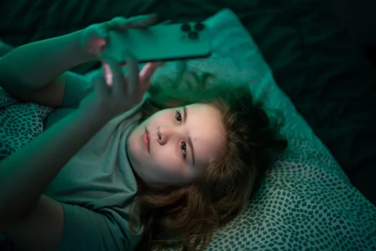 Teenage girl lying in bed in the dark, her face lit by the smartphone she is holding above her