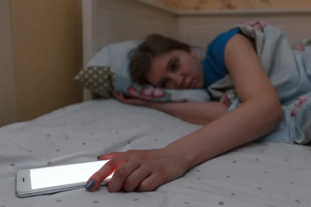 Teenage girl lying in bed in a dark room, reaching for a glowing smartphone