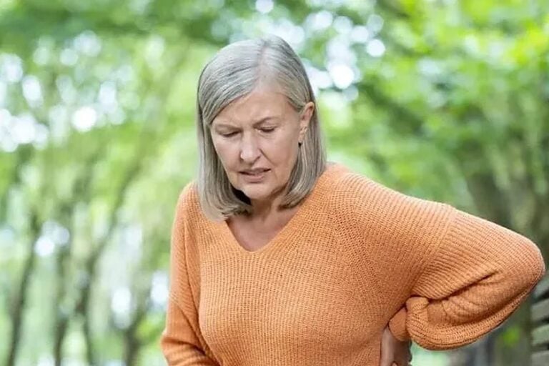 Older woman in an orange sweater holding her side in pain outdoors