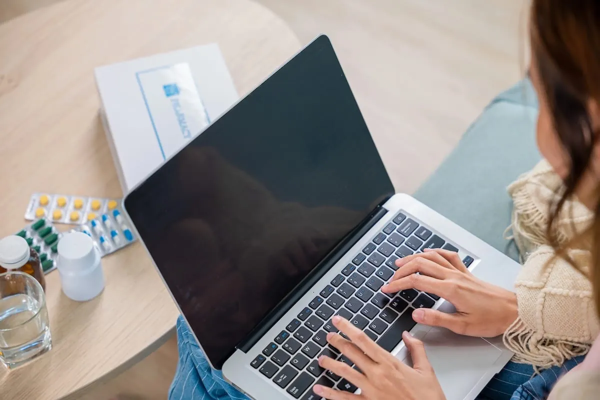 Woman typing on a laptop beside blister packs of pills, medicine bottles, a glass of water and a pharmacy box